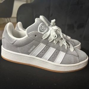 Grå Adidas Campus sneakers - Säljer ett par snygga grå Adidas Campus sneakers i mocka med vita detaljer och snörning. De har den klassiska tre-randiga designen på sidorna och en bekväm gummisula. Perfekta för vardagsbruk och ger en stilren look. Också perfekt inför julen som julklapp!