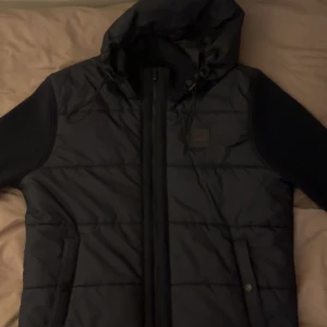 Mörkblå cardigan jacka med huva - Säljer en snygg mörkblå  cardigan quiltad jacka från Boss. Den har en dragkedja framtill och en praktisk huva. Ärmarna är stickade vilket ger en cool kontrast till det quiltade materialet. Perfekt för höst och vår! Pris ej hugget i sten!