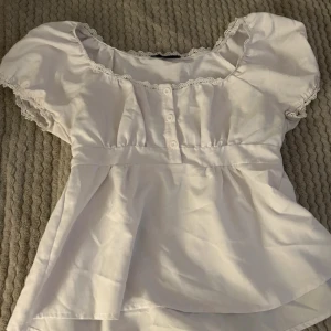 Vit blus med spetsdetaljer - Söt vit blus med spetsdetaljer runt halsen och ärmsluten. Blusen har korta ärmar och knappar framtill.  Perfekt för vår och sommar