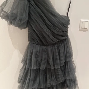 Grå klänning från By Malina - Säljer en elegant grå klänning från By Malina. Den har en oneshoulder-design med volanger och en luftig, flerdelad kjol. Perfekt för festliga tillfällen!