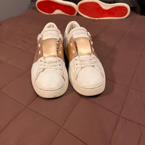 Valentino sneakers i vitt och guld - Säljer ett par snygga vita sneakers från Valentino med gulddetaljer. Skorna har en bred guldfärgad rem över snörningen och coola nitar på hälen. Perfekta för att ge din outfit en lyxig touch! De är i bra skick med lite små defekter som ej syns vid användning och storleken är 41 men funkar båda för 40 med inkluderade sulan och som 42 med tunnare sula nypris ligger på 5000-6000. Kom gärna med bud eller bytesförslag 