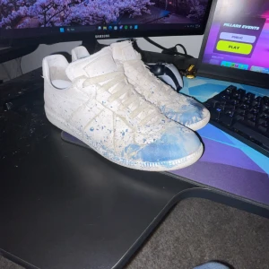 Vita sneakers från Maison Margiela - Unika vita sneakers från Maison Margiela med blå färgstänk och snörning. Skorna har en distinkt design med en robust sula och är perfekta för den som vill sticka ut. Lägg bud. Box och dustbag medkommer vid köp