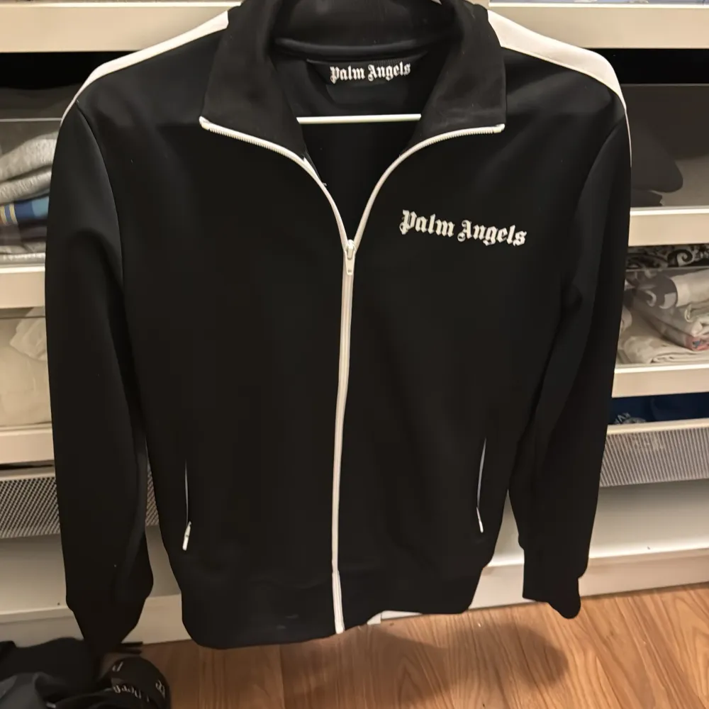 Snygg svart track jacket från Palm Angels med vita detaljer och dragkedja. Jackan har en klassisk design med logotyp på bröstet och vita ränder längs ärmarna. Perfekt för en sportig look.. Hoodies.