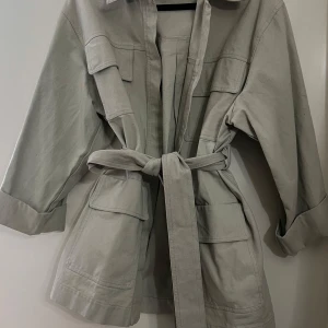 Mintgrön jeansjacka med knytband - Snygg mintgrön jeansjacka med knytband i midjan och stora fickor framtill. Jackan har en klassisk krage och trekvartsärmar, perfekt för vår och höst. En stilren och mångsidig jacka för alla tillfällen. Köpt på GinaTricot storlek L men passar m/s om man tycker om lite mer oversized stil.