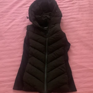 Svart dunväst från Zara - Snygg svart dunväst från Zara med huva och dragkedja. Perfekt för kyligare dagar och ger en stilren look. Västen har ett quiltat mönster som ger extra värme och komfort.