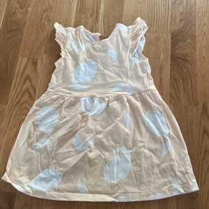 Beige och vit klänning från H&M - Söt beige klänning från H&M med stora vita detaljer och volanger vid ärmarna. Perfekt för små barn som vill ha något bekvämt och stilrent. Klänningen har en rund hals och är ärmlös, vilket gör den idealisk för varmare dagar.