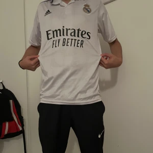 Vit Real Madrid fotbollströja - Säljer en vit Real Madrid fotbollströja från Adidas med korta ärmar. Tröjan har texten 'Emirates Fly Better' på framsidan och 'Benzema 9' på baksidan. Perfekt för fans av laget!