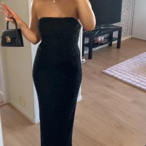 Svart glittrig axelbandslös klänning - Säljer en elegant svart glittrig klänning som är axelbandslös och figurnära. Perfekt för festliga tillfällen! Klänningen är lång och har en stilren design som verkligen fångar uppmärksamheten. Passar perfekt för nyår eller andra speciella tillfällen. 🖤✨