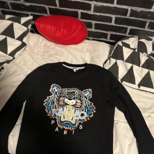 Svart tröja med tigerbroderi från Kenzo - Säljer en svart tröja från Kenzo med ett coolt tigerbroderi i flera färger på framsidan. Tröjan har långa ärmar och rund halsringning. Perfekt för dig som vill ha en stilren men ändå iögonfallande look. Pris kan diskuteras 