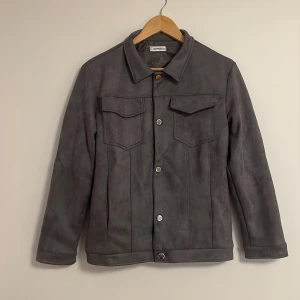 Grå mockajacka från ZULA STOCKHOLM - Snygg grå mockajacka med knappar framtill och två bröstfickor. Jackan har en klassisk krage och långa ärmar, perfekt för en stilren look.