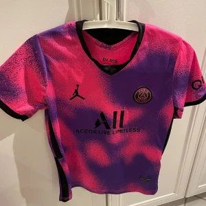 PSG fotbollströja med Mbappé 7 - Säljer en PSG fotbollströja i lila och rosa med Mbappé 7 på ryggen.  Tröjan är kortärmad och har en cool design med svart text och detaljer. Perfekt för fans av både PSG och Mbappé. (Storlek 11-12år) 