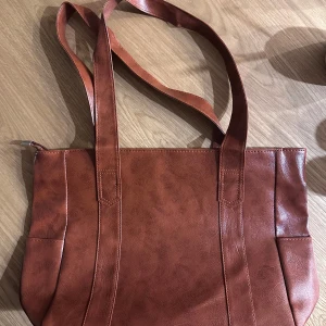Handväska - En helt ny och oanvänd brun axelväska. Den har en praktisk dragkedja upptill och gott om plats inuti för alla dina grejer. Perfekt för vardagsbruk eller när du behöver ha med dig lite extra saker, som skolan eller resa!! Väskan har långa remmar så den är lätt att bära över axeln. Mått: 42cmx27cmx11cm
