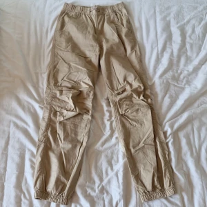 Beige byxor från Lindex - Säljer ett par bekväma beige cargobyxor från Lindex. De har en elastisk midja med snörning för extra komfort och justerbar passform. Byxorna är perfekta för vardagsbruk och har en avslappnad stil med resår vid bensluten.