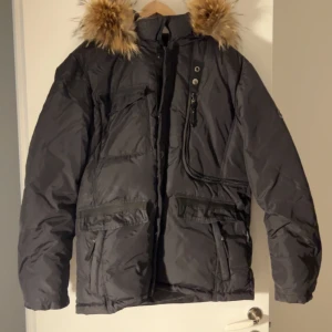 Svart vinterjacka med pälsdetaljer - Säljer en svart vinterjacka från Hollies med snygga pälsdetaljer på huvan. Jackan har flera praktiska fickor och en dragkedja framtill. Perfekt för kalla vinterdagar! Kvalite 9/10! Nypris 4000kr. Mitt pris 899kr.