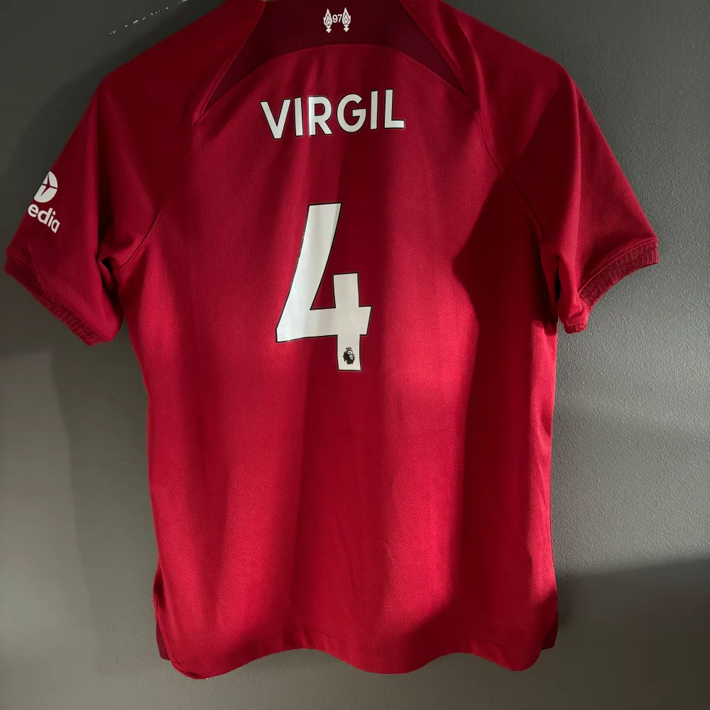 Säljer förra årets Liverpooltröja från Nike i storlek 158-170cm. Tröjan har korta ärmar och är i Dri-FIT material för bra komfort. Den har Liverpool FC-loggan och sponsortryck på framsidan, samt 'VIRGIL' och nummer 4 på baksidan. Nypris: 999kr. T-shirts.