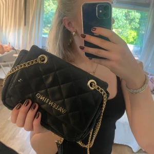 Svart quiltad handväska från Valentino - Säljer en snygg svart handväska från Valentino. Väskan är quiltad och har en elegant design med guldfärgade detaljer. Den har en kedja som axelrem och en liten handtag för att bära den i handen. Perfekt för både vardag och fest!