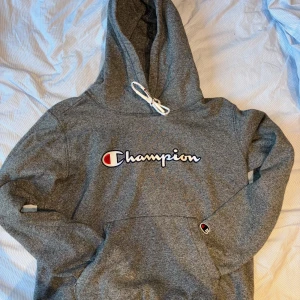Grå hoodie från Champion - Säljer en snygg grå hoodie från Champion i storlek S. Den har en klassisk Champion-logga på bröstet och en liten logga på ärmen. Hoodien är perfekt för höst och vinter med sin mysiga och varma känsla. Den har en justerbar huva med vita snören och en stor ficka framtill. Perfekt för chill dagar eller en casual look!