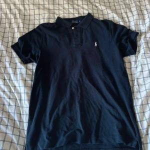 Svart pikétröja från Ralph Lauren - Säljer en klassisk svart pikétröja från Ralph Lauren. Den har en liten broderad logga på bröstet och är i storlek M. Perfekt för både vardag och lite finare tillfällen. Tröjan är i bra skick och redo för en ny ägare!