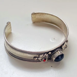 Silverarmband med dekorativa stenar - Säljer ett snyggt silverfärgat armband med en stor svart sten i mitten och en liten röd sten bredvid. Saknar en sten. Okänd metall. Armbandet har fina detaljer med blommönster och små pärlor. Perfekt för att ge en elegant touch till vilken outfit som helst!