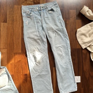 Ljusblå jeansbyxor med slitningar - Säljer ett par ljusblå jeansbyxor med coola slitningar på låren. De har en straight passform och är perfekta för en avslappnad stil. Byxorna har klassiska fem fickor och stängs med dragkedja och knapp. Perfekta för vår och sommar!