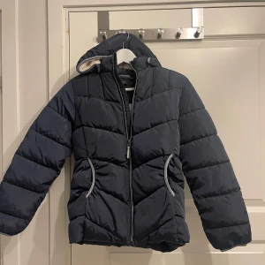 Mörkblå dunjacka från Name It - Säljer en mörkblå dunjacka från Name It i mycket bra skick. Jackan har en mysig fleecefoder på insidan och en praktisk dragkedja framtill. Perfekt för kalla vinterdagar med sin varma och quiltade design. Den har långa ärmar och en huva för extra skydd mot kylan.