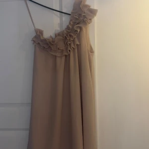 Beige/rosa oneshoulder klänning - Säljer en superfin beige/rosa oneshoulder klänning med volangdetaljer. Perfekt för sommarens fester eller en elegant middag. Klänningen har en luftig passform och är gjord i ett lätt material som faller snyggt. Ett måste i garderoben för den som vill ha något stilrent men ändå speciellt! (Använd vid ett tillfälle)