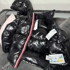 Svart glansig dunjacka från Moncler - Säljer en snygg svart glansig dunjacka från Moncler. Jackan har en cool röd och vit rand längs dragkedjan och en praktisk huva. Perfekt för kalla dagar med sin pufferstil och varma fyllning. Den har en klassisk Moncler-logga på bröstet. Passar perfekt för vintern och hösten!
