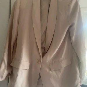 Beige kavaj från NLY Trend - Säljer en stilren beige kavaj från NLY Trend i storlek 38. Kavajen har en klassisk design med en knapp framtill och långa ärmar. Perfekt för både vardag och festliga tillfällen. Passar bra till höst och vår! Defekt baktill längst ned, vet ej om det går att få bort. 