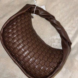 Brun flätad axelväska - Snygg brun axelväska i fakeskinn med flätad design. Väskan har en unik tvinnad axelrem och dragkedja upptill. Perfekt för både vardag och festliga tillfällen. Den är i helt nytt skick och redo att användas direkt!