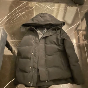 Svart vinterjacka från Canada Goose - Säljer en svart vinterjacka från Canada Goose i mycket bra skick. Jackan har en huva och stängs med dragkedja och knappar. Perfekt för kalla vinterdagar. Den är varm och stilren, idealisk för både vardag och utomhusaktiviteter. Pris kan diskuteras 