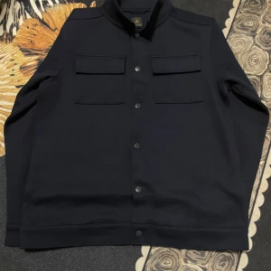 Svart kappa från Next - Säljer en stilren svart jacka från Next i mycket bra skick. Jackan har en klassisk design med två bröstfickor och knappar framtill. Den är tillverkad av återvunnen polyester, vilket gör den både hållbar och miljövänlig. Perfekt för höst och vår!