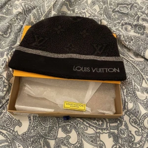 Svart Louis Vuitton Mössa - Säljer en snygg svart mössa från Louis Vuitton med grå detaljer och logotyp. Perfekt för att hålla sig varm med stil. Kommer i originalförpackning med dustbag. Passar både till vardags och festliga tillfällen.