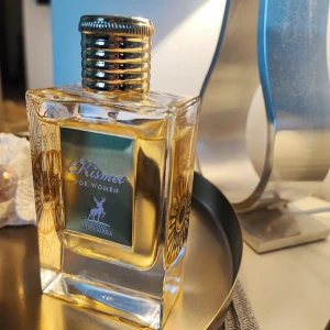 Kismet for Women från Maison Alhambra - Säljer en elegant parfymflaska av Kismet for Women från Maison Alhambra. Flaskan är fyrkantig med en gyllene etikett och lock. Doften har toppnoter av aprikos, ros och jasmin, mellannoter av tuberos och narciss, samt basnoter av amber och trä. Perfekt för den som älskar blommiga och fruktiga dofter.