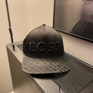  Hugo boss keps - Snygg svart keps från Boss med ett coolt mönster på skärmen och broderad text framtill. Perfekt för att ge din outfit en stilren touch. Kepsen har justerbar passform och är i mycket bra skick.