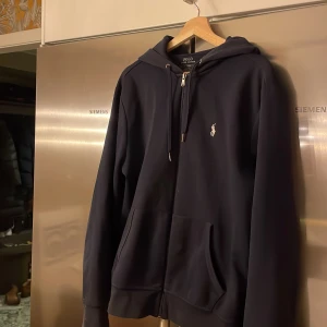 Blå hoodie från Polo Ralph Lauren - Säljer en stilren blå hoodie från Polo Ralph Lauren i storlek L. Den har en klassisk design med dragkedja och den ikoniska loggan på bröstet. Perfekt för både vardag och chill. Grymt skön och passar till allt! 🖤