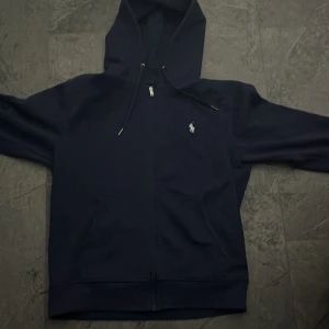 Mörkblå hoodie med dragkedja - Säljer en snygg mörkblå hoodie från Polo Ralph Lauren. Den har en dragkedja framtill och en liten broderad logga på bröstet. Perfekt för höst och vinter, och den är superbekväm att ha på sig. Passar både till vardags och chillkvällar.