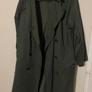 Grön trenchcoat från Zara - Säljer en snygg grön trenchcoat från Zara i storlek XS. Den har en klassisk design med knappar framtill och en stilren look. Perfekt för höst och vår när du vill ha något lätt men ändå skyddande. Passar både till vardags och lite finare tillfällen.