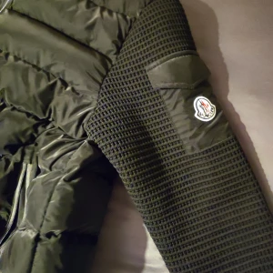 Svart quiltad jacka - Säljer en snygg svart quiltad jacka. Den har långa ärmar med ett coolt mönster och en dragkedja framtill. Perfekt för höst och vinter! Jackan är i mycket bra skick och har en stilren design med Moncler-loggan på ärmen.