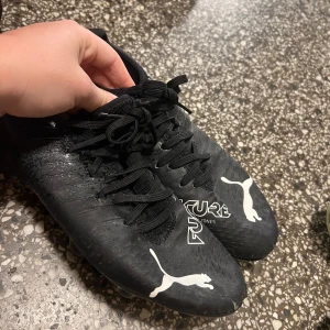 Svarta fotbollsskor från Puma - Säljer ett par svarta fotbollsskor från Puma i bra skick. Skorna har snörning och är perfekta för fotbollsträning. De har en stilren design med vita Puma-loggor och dobbar för bra grepp på planen.