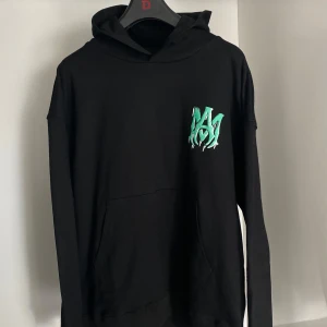 Amiri Watercolor Hoodie - Säljer en svart hoodie från Amiri i nyskick! Den är helt ny och oanvänd med alla etiketter fortfarande kvar. Den har en cool grön logga på bröstet och AMIRI skrivet stort på ryggen. Perfekt för höst och vinter, och den är superbekväm med en känguruficka framtill.