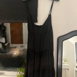 Svart långklänning med volanger - Säljer en elegant svart långklänning med smala axelband och volangdetaljer. Perfekt för festliga tillfällen eller en kväll ute. Klänningen har en luftig passform. Aldrig använt