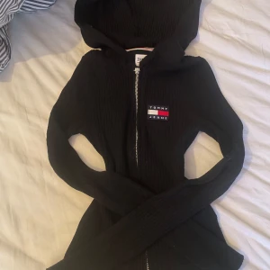 Svart tunn kofta från Tommy Jeans - Säljer en svart tunn kofta från Tommy Jeans i ribbat material. Den har en dragkedja framtill och en klassisk logga på bröstet. Perfekt för höst och vinter, och den är i mycket bra skick. Säljer för att den inte kommer till användning❤️