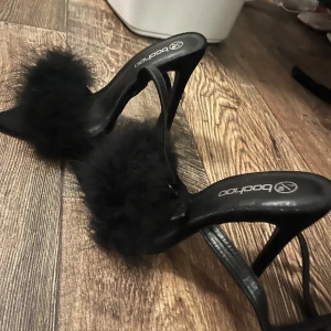 Svarta klackskor med pälsdetalj från Boohoo - Snygga svarta klackskor från Boohoo med en fluffig pälsdetalj över foten. De har en spetsig tå och en elegant klack som passar perfekt för festliga tillfällen. Skorna är i bra skick och redo att användas för att ge din outfit en extra touch av glamour.