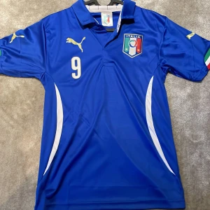 Blå fotbollströja från Puma - Säljer en blå kortärmad fotbollströja från Puma med Italiens landslagsemblem på bröstet. Tröjan har nummer 9 och namn på baksidan. Perfekt för fotbollsfans! 🏆