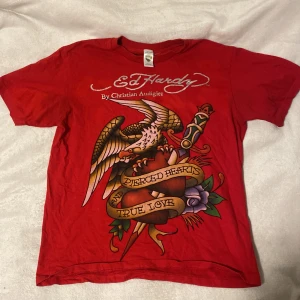 Röd Ed Hardy t-shirt med tryck - Säljer en cool röd t-shirt från Ed Hardy med ett stort tryck av en örn och ett hjärta med texten 'Pierced Hearts True Love'. T-shirten är kortärmad och har märkets logga både fram och bak. Använt den Max 2 gånger, jätte bra skick! Skulle säga att den är S/M, den är lite mer åt det oversized hållet.