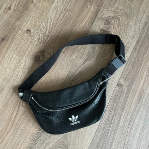 Svart magväska från Adidas - Säljer en snygg svart magväska från Adidas i skinnmaterial. Den har en justerbar rem och en praktisk dragkedja framtill. Perfekt för att ha med sig det viktigaste på språng. Passar både till vardags och fest!