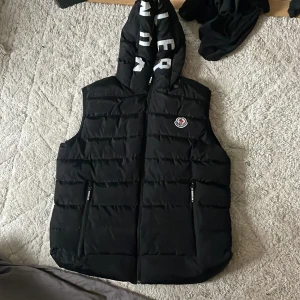 Svart dunväst från Moncler - Säljer en snygg svart dunväst från Moncler i storlek L. Västen har en quiltad design med dragkedja och en huva med Moncler-logga. Perfekt för höst och vinter, håller dig varm och stilren. Passar både till vardags och festliga tillfällen.