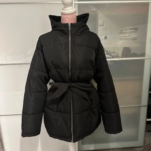 Svart pufferjacka med bälte - Säljer en svart pufferjacka med bälte i midjan från Nelly. Jackan har en dragkedja framtill och långa ärmar. Perfekt för kalla dagar när du vill hålla dig varm och snygg. Passar både till vardags och lite finare tillfällen. Även helt ny tvättad, säljer då jag köpte den på Vinted men den var för stor. 