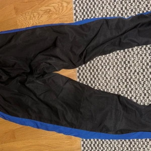 Svarta träningsbyxor från Lacoste Sport - Säljer ett par svarta träningsbyxor från Lacoste Sport i storlek US S. Byxorna har blå detaljer längs sidorna och är perfekta för träning. De är i bra skick och har en bekväm passform. Perfekta för både gymmet och avslappnade dagar.