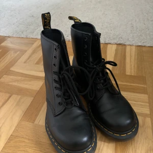 Svarta boots från Dr. Martens - Säljer ett par klassiska svarta Dr. Martens 1460 boots i skinn. De har den ikoniska gula sömmen och snörning framtill. Perfekta för både höst och vinter, och de är i mycket bra skick. Passar till nästan alla outfits och är superbekväma att gå i. Perfekta för dig som vill ha en tidlös stil!